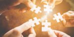 Hands holding white puzzle pieces, coming together as a team, symbolizing collaboration and adaptive leadership in business