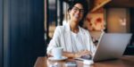 Smiling entrepreneur working on laptop in a café, representing business coaching success stories and the real results entrepreneurs achieve through coaching.