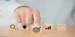 A hand points at a wooden block with a target icon, symbolizing high ticket sales success. Other blocks display icons of a magnifying glass, light bulb, graph, and clock, representing analysis, ideas, growth metrics, and time management.