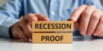 Hands stacking wooden blocks labeled recession proof, representing strategies for building recession proof businesses in 2026