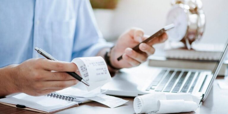 A person using a smartphone and holding receipts while working on a laptop, organizing financial documents to understand revenue vs profit.